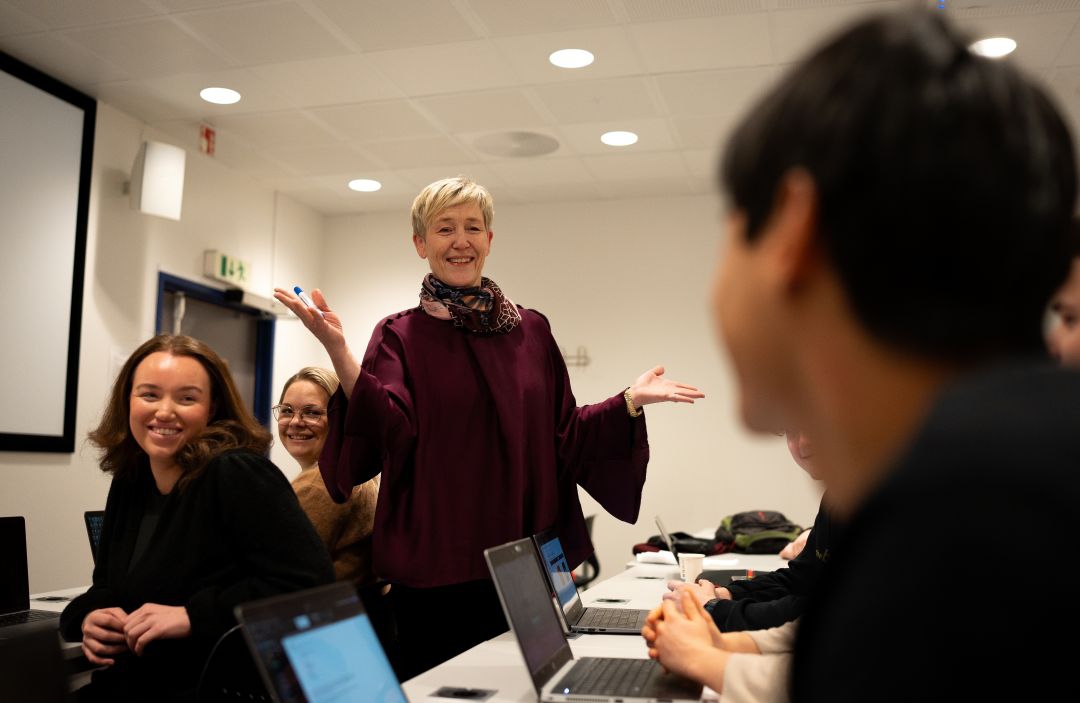 Foreleser står midt i et klasserom og forklarer for studentene. Bruker armene til å vise engasjement.