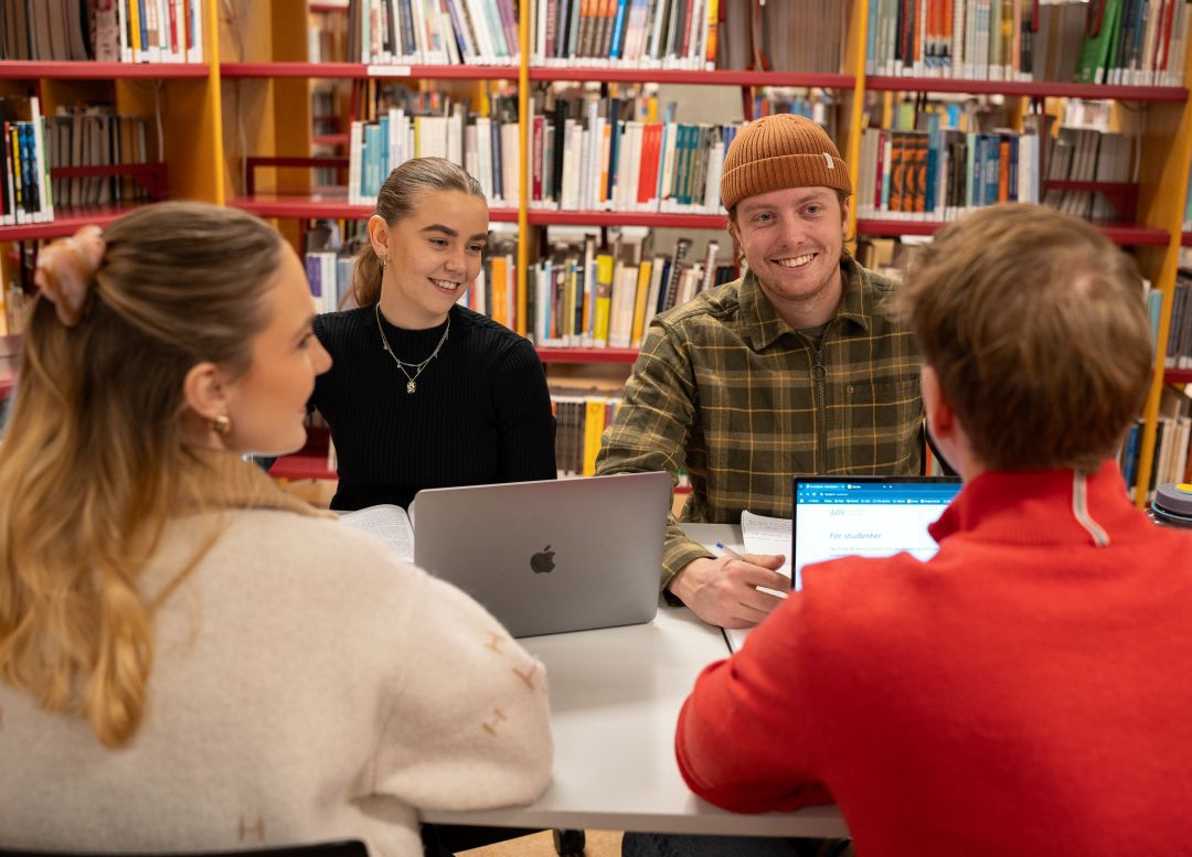 Fire studenter sitter å jobber ved et bord i biblioteket på campus. Mange bøker i bakgrunn.