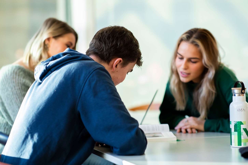 Tre sittende studenter snakker sammen mens de kikker i en bok.