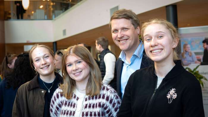 Tre ungdommer og utviklingsminister smiler til kamera, folk i bakgrunnen.