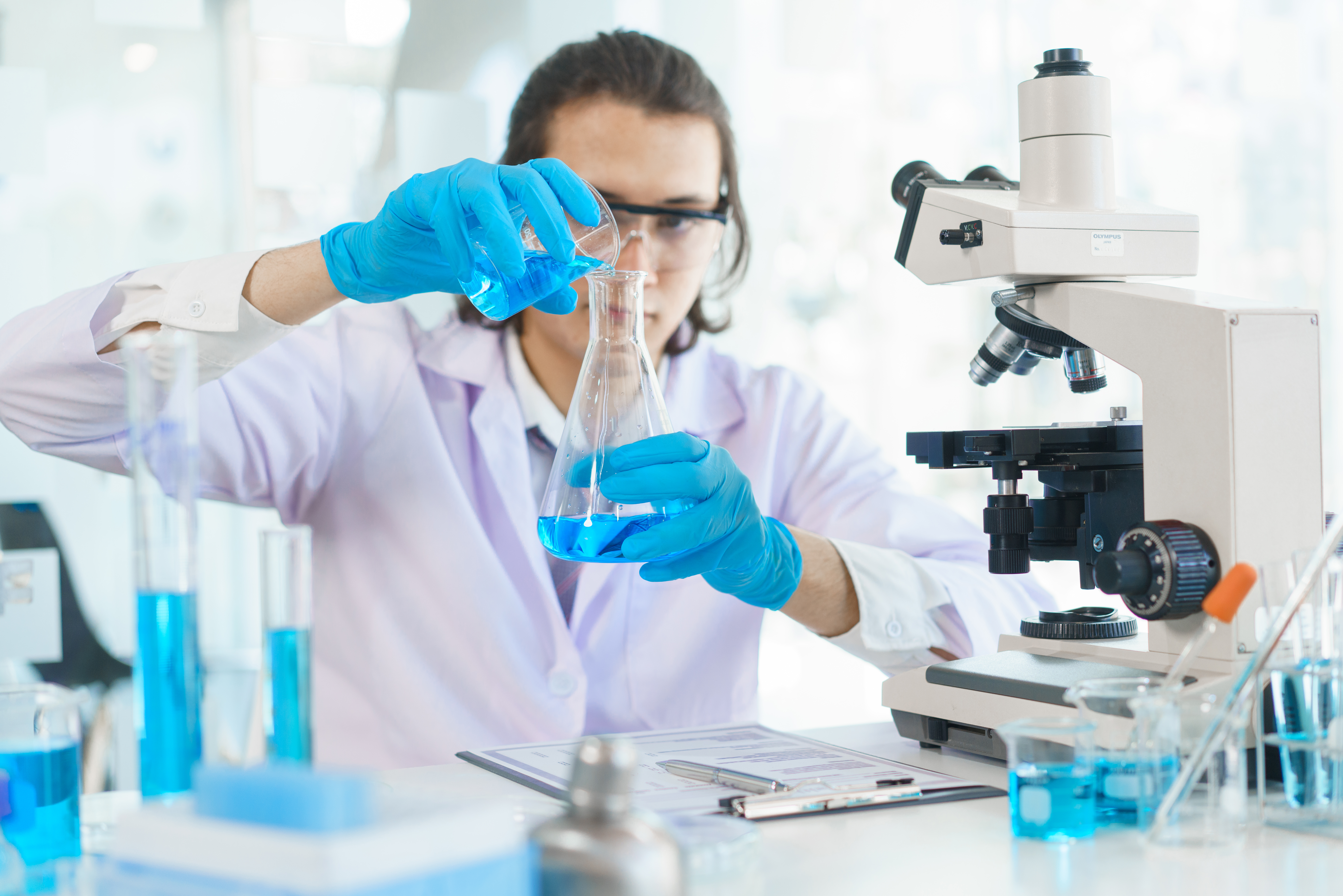 En forsker sitter på et laboratorium med blå hansker og vernebriller. Foto: Colourbox