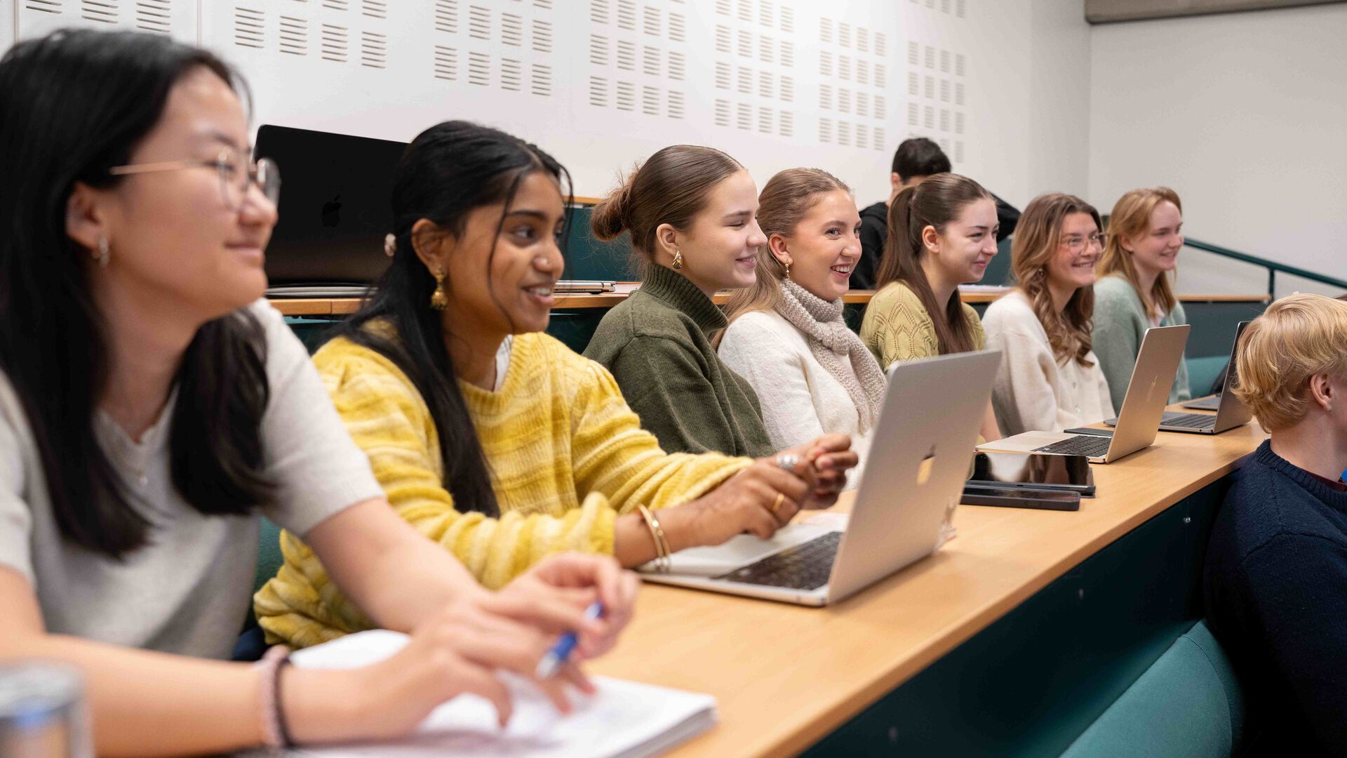 Sju blide studenter sitter på rekke i en forelesningssal, noen med pc, andre med penn og notatbok. Studenter skimtes også på rekkene foran og bak.