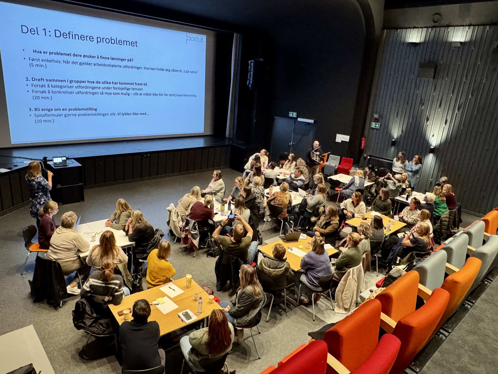 Oversiktsbilde over en sal med ca 50 mennesker som sitter rundt bord på gulvet og følger med på en presentasjon på storskjerm. Tittel på informasjonen på skjermen er Del 1: Definere problemet
