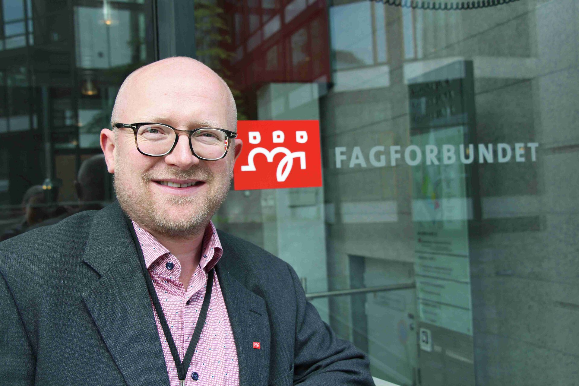 Finstad i dressjakke og rød skorte foran et vindu med skriften FAGFORBUNDET og rød logo
