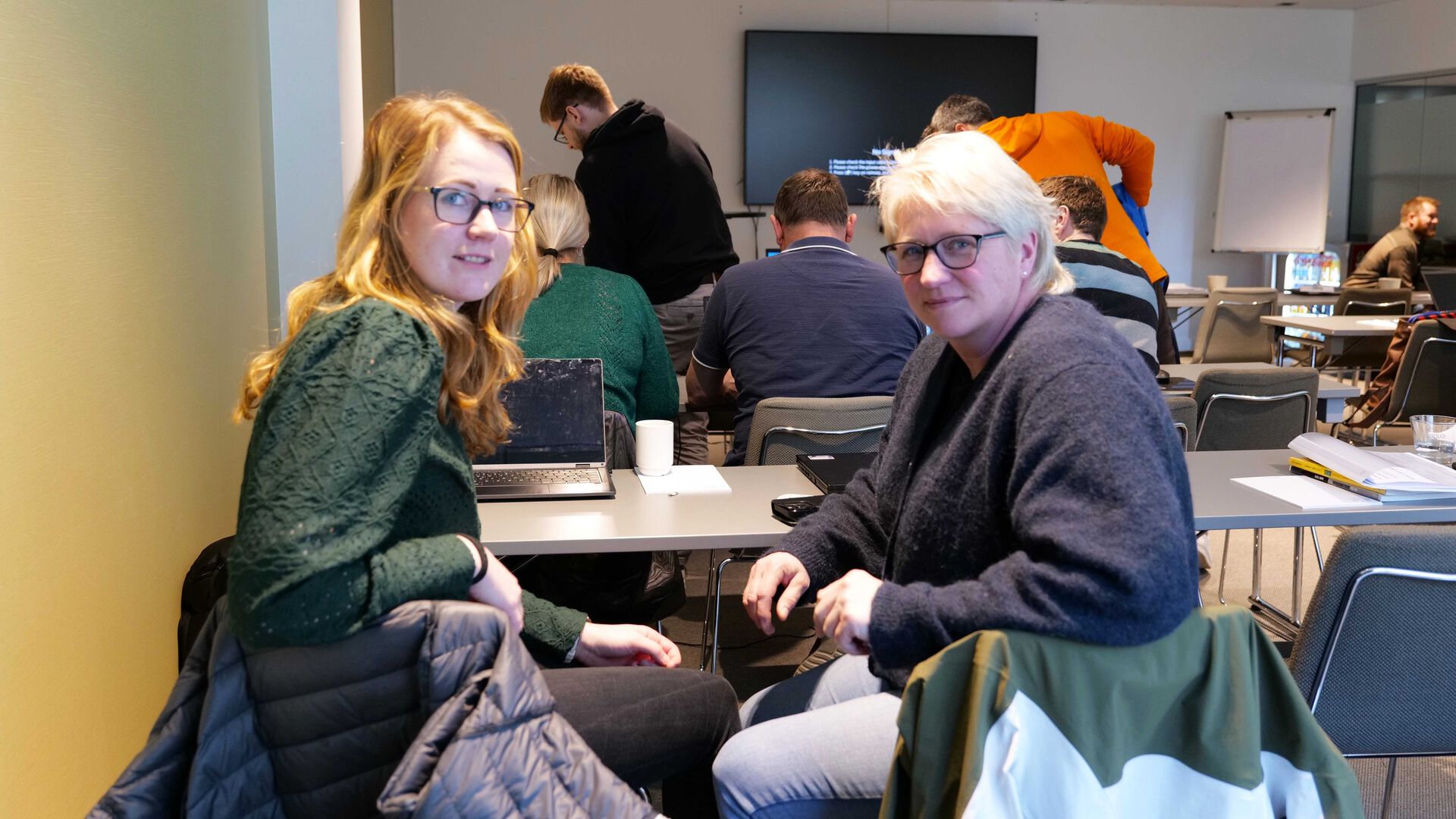 Datter og mor sitter ved siden av hverandre i klasserommet, snur seg og ser på fotografen. Storskjerm og flere studenter i bakgrunnen.
