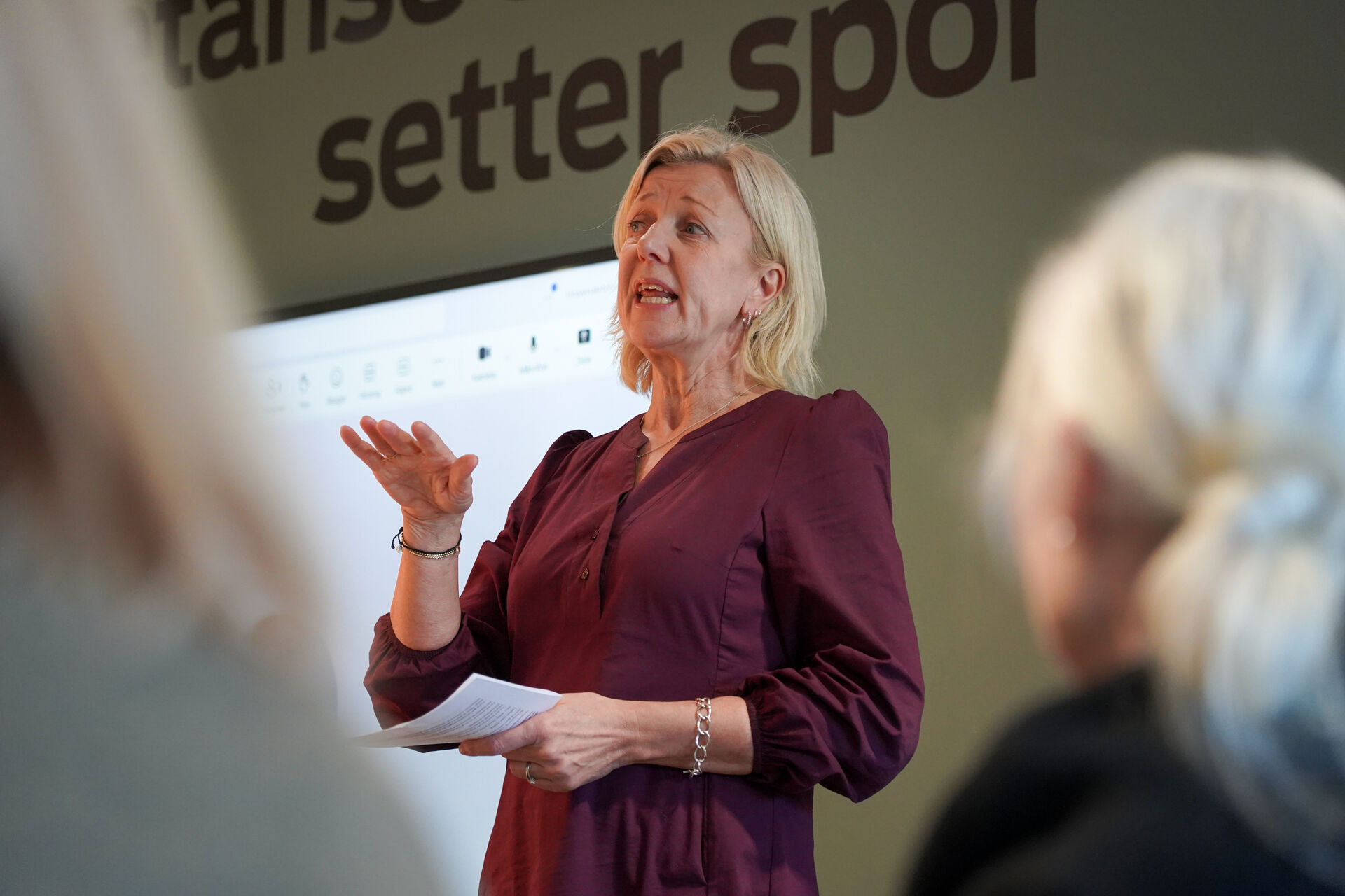 Dame foran en mosegrønn vegg med teksten "Setter spor" holder tale