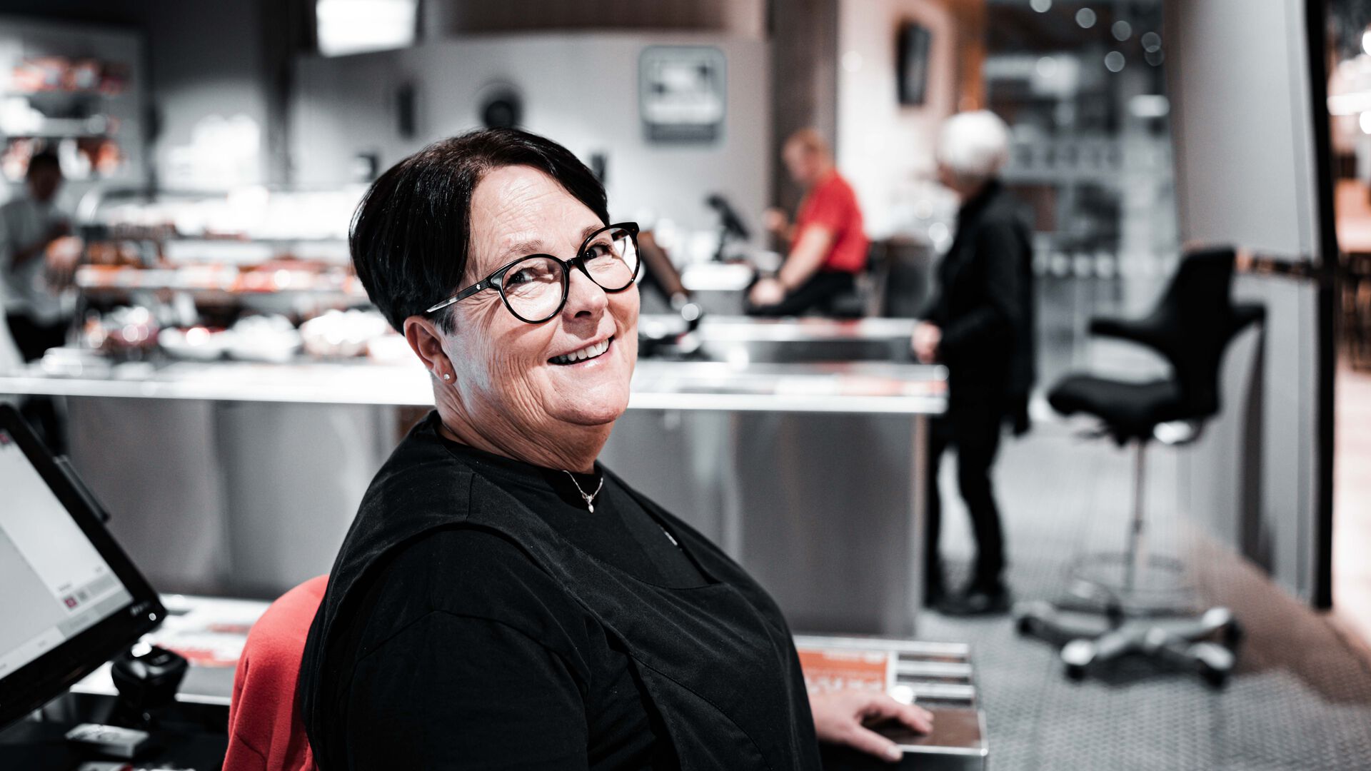 Tove Svisdal sitter i kassa i kantina på Lillehammer. Serveringsdisk og folk diffust i bakgrunnen.