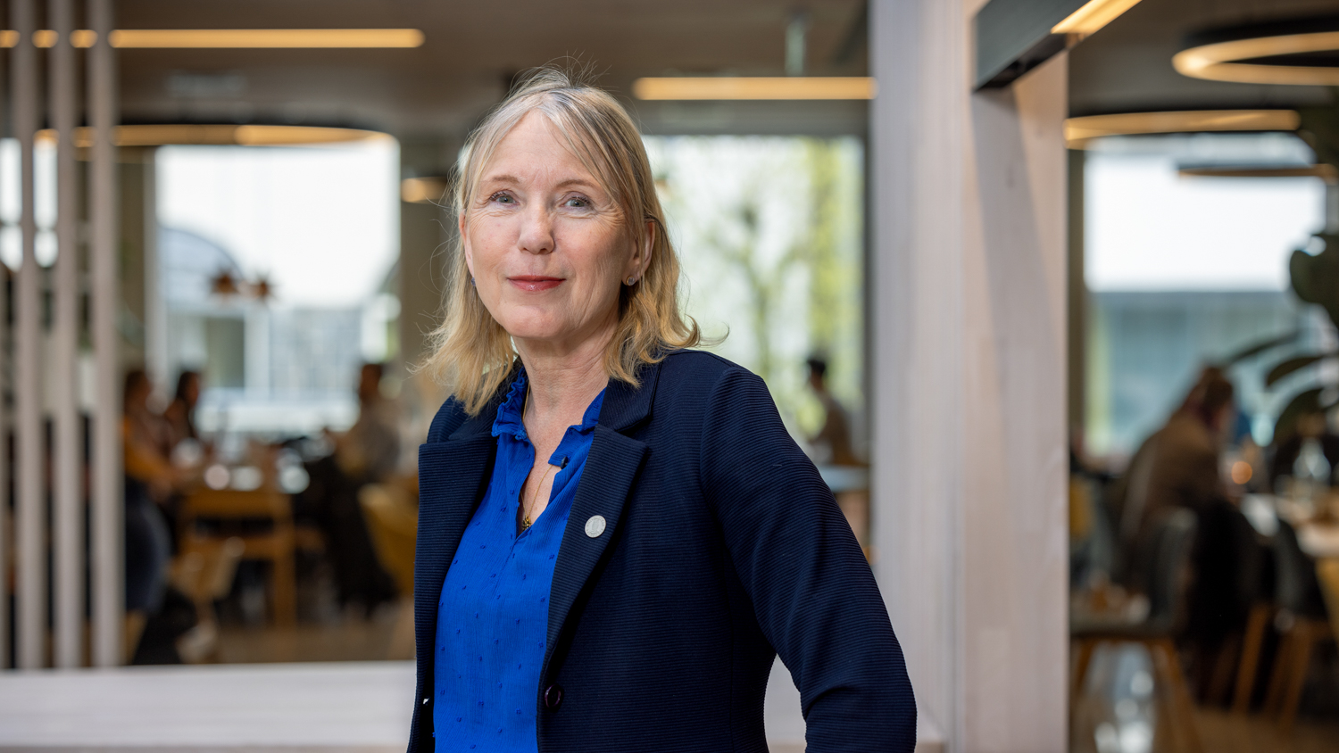 Rektor på Universitetet i Bergen med blå skjorte og mørkeblå jakke inne på campus med noen store vinduer i bakgrunnen 