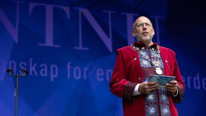 Rektor på NTNU, Tor Grande, med festantrekk mot blå bakgrunn der det står NTNU med store bokstaver