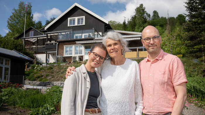 Tre personer står sammen foran en blomsterhage og et svart hus i bakgrunnen