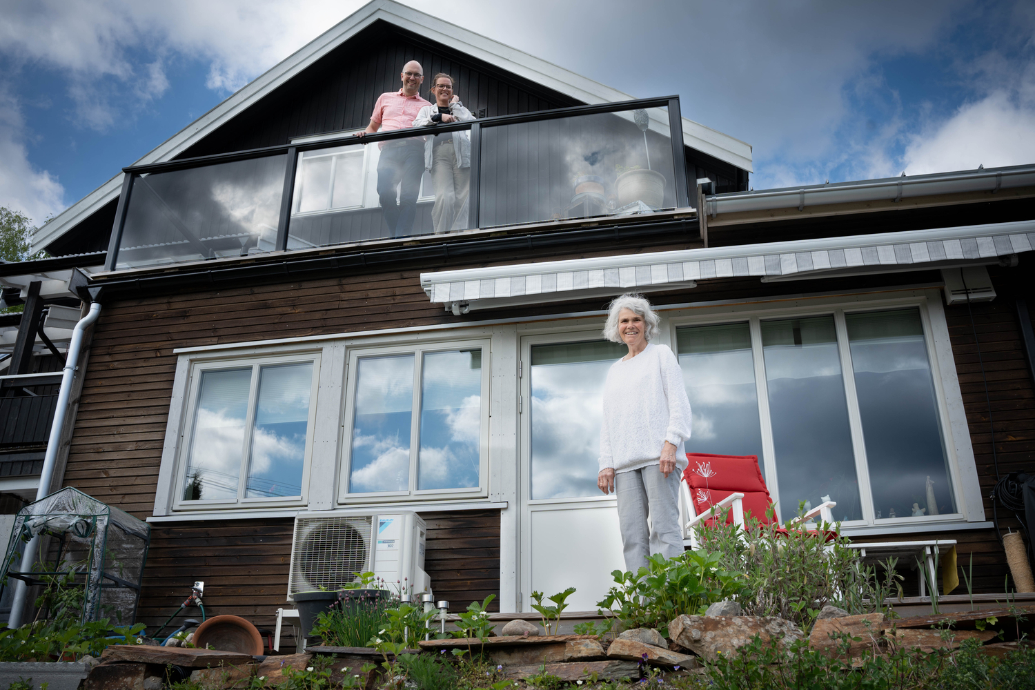 Gammel dame foran et hus med et yngre par stående på en veranda over henne