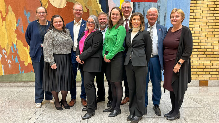 Ni stortingspolitikere sammen i Stortingets vandrehall foran et vevd teppe og en lys murvegg