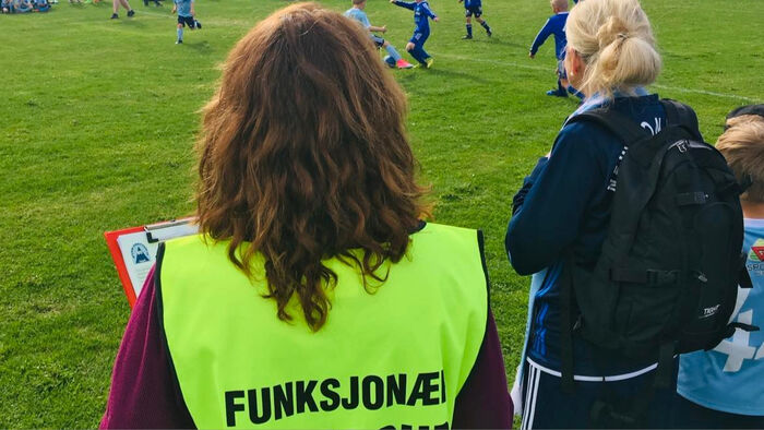 dame med gull vest og funksjonær på ryggen ser på barnefotballkamp.