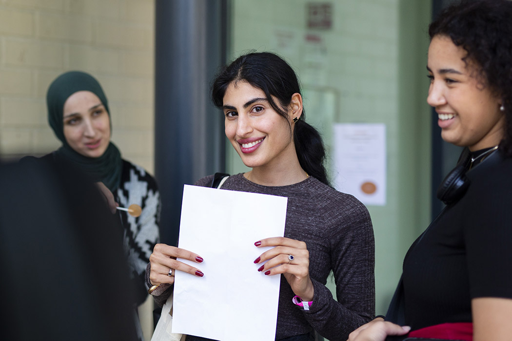 Tre glade studenter snakker sammen, og det midterste ser rett inn i kameraet med et stort smil og et hvitt ark i hendene