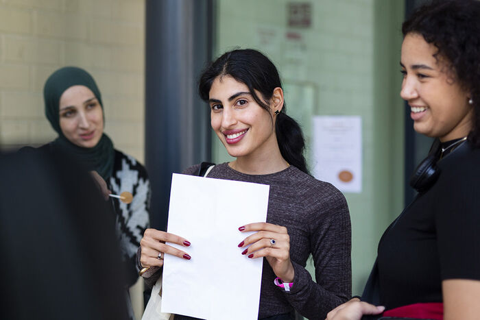 Tre glade studenter snakker sammen, og det midterste ser rett inn i kameraet med et stort smil og et hvitt ark i hendene