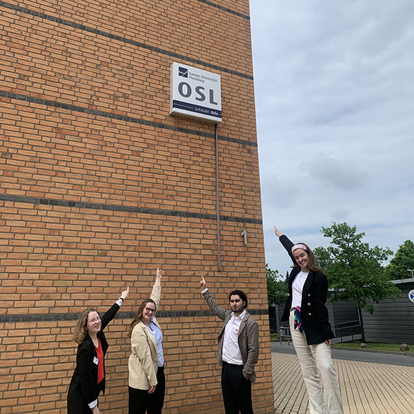 Fire studenter står utenfor et stort murbygg og peker på et skilt som sier OSL