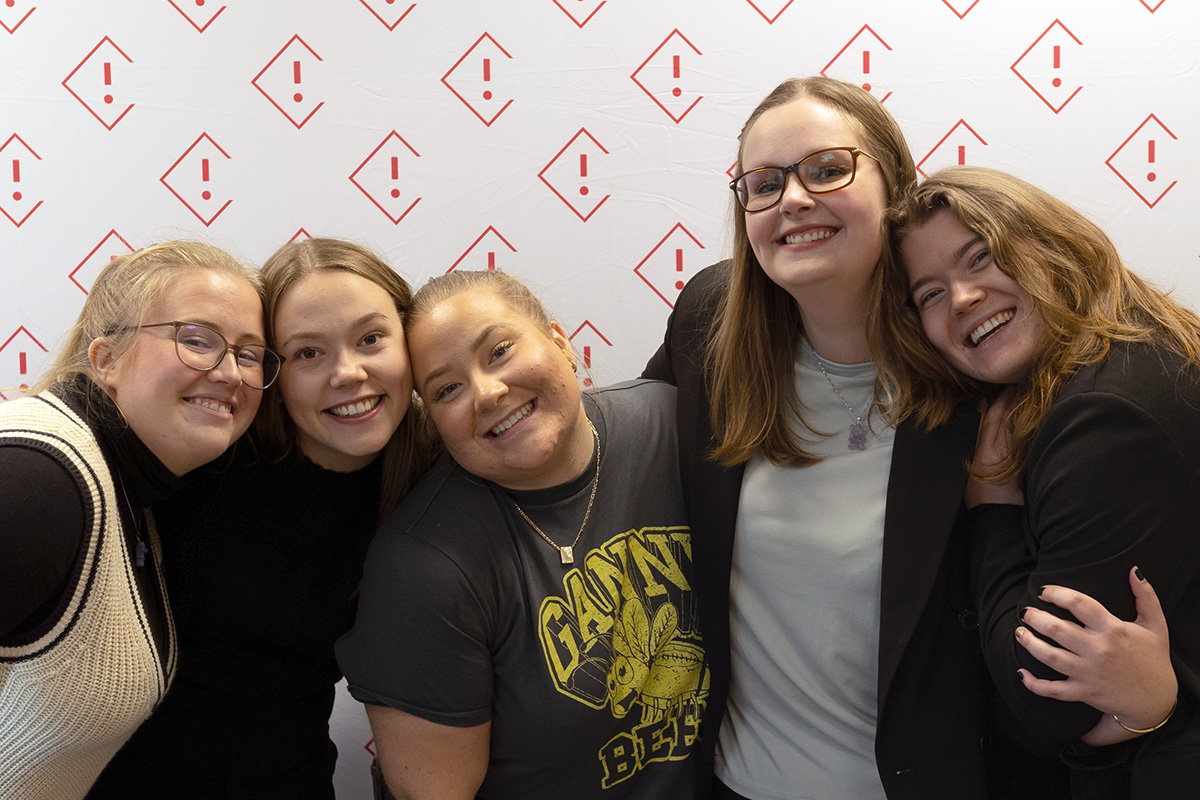 Fem glade kvinner smiler inn i kamera mens de holder rundt hverandre foran en vegg med StInn-logoer