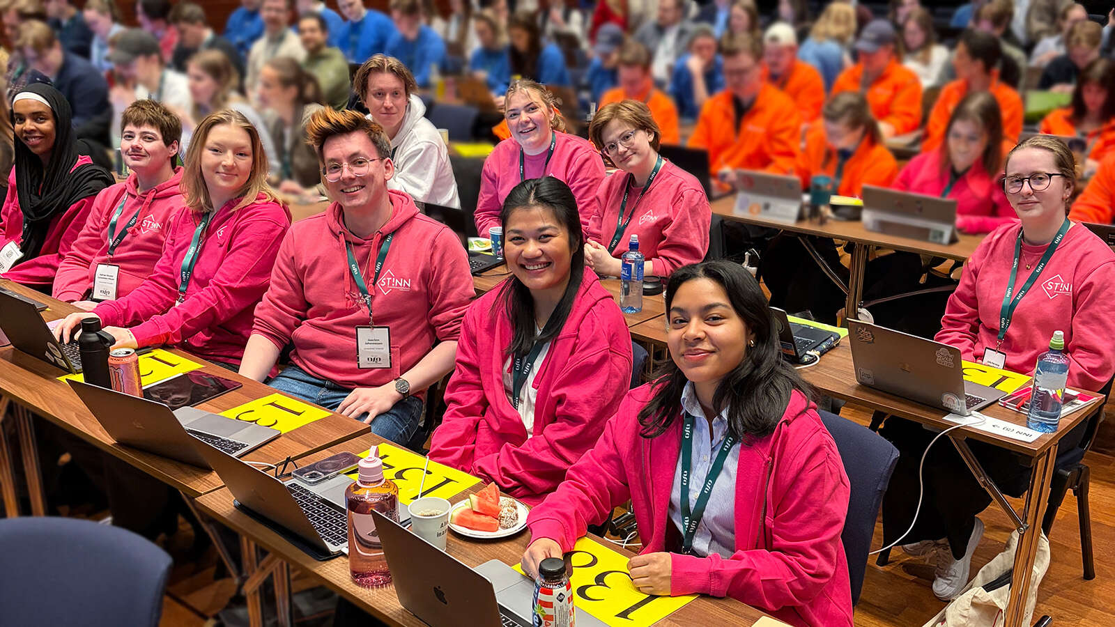 Delegasjonen fra StInn sitter ved plassene sine i landsmøtesalen, som er fylt med delegater. Alle fra StInn har rosa hettegensere og ser mot kameraet med et smil.