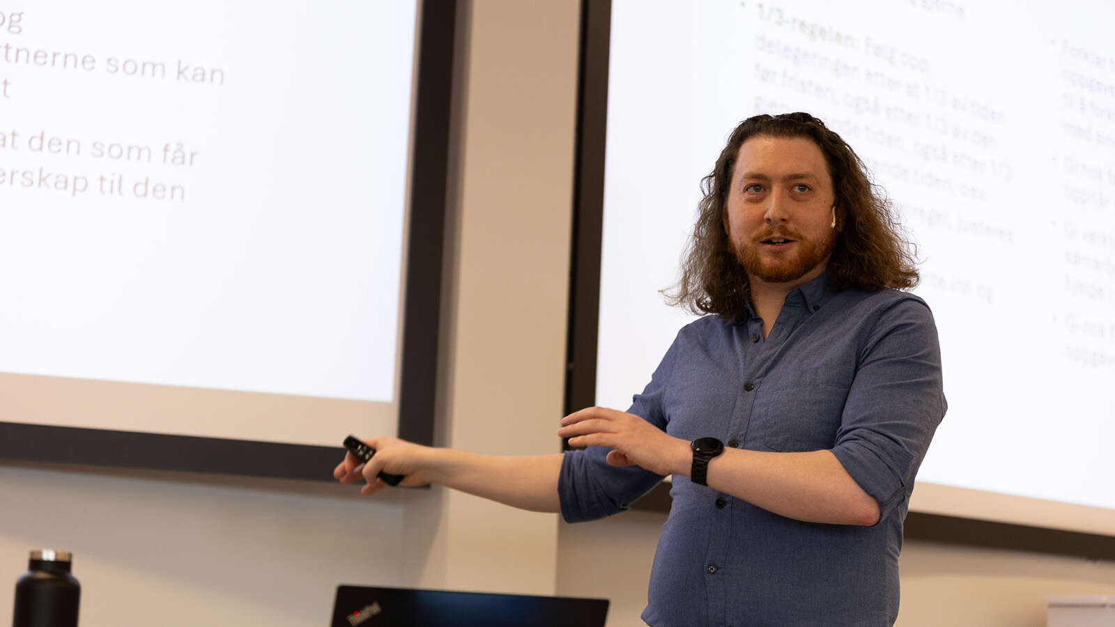 En mann gestikulerer med en fjernkontroll i hånden mens han presenterer en PowerPoint-presentasjon