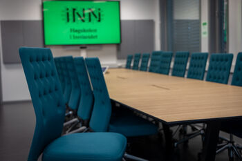 Et formelt møterom hvor identiske kontorstoler er satt pent inntil et stort konferansebord. Ved enden av bordet er en stor skjerm hvor Høgskolen i Innlandets logo blir vist. Mesteparten av møterommet er utenfor fokus, bortsett fra en ledig stol som er trukket fra bordet, klar for at noen skal sette seg ned på den.