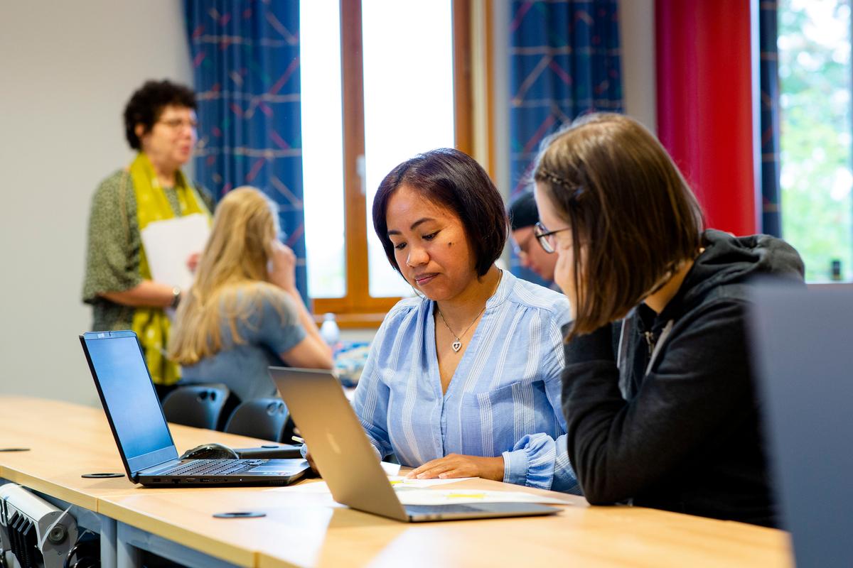 To kvinnelige studenter i klasserom men laptoper oppe. Foreleser er å se i bakgrunn.