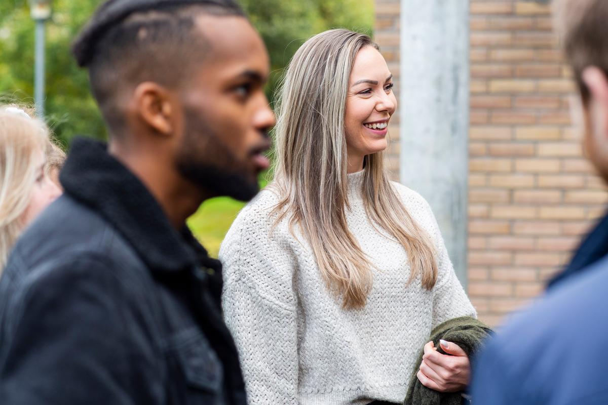Studenter som står å snakker utenfor et høgskolebygg. En mørk mann i fokus og en dame med langt blondt hår. Hun har jakken under armen.