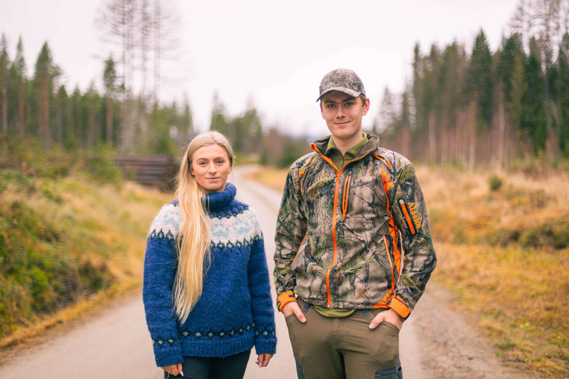 Skogstudenter med friluftsklær står å ser i kamera. De står på en grusveg i skogen. Ser skog og tømmer på begge sider. Høstvær.