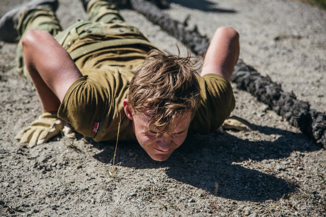 Soldat som ligger ned i sanden og prøver komme seg opp med armene. Et tau ved siden av.