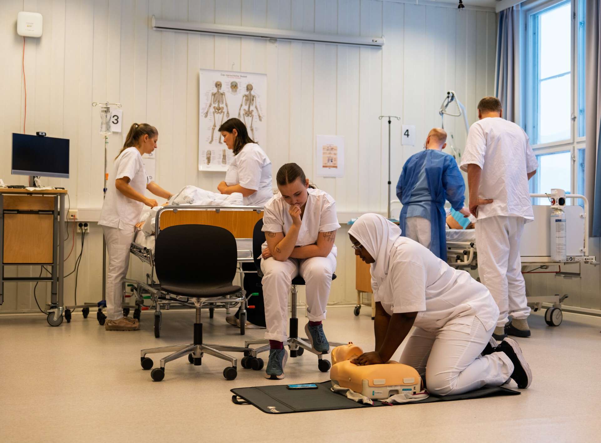 Sykepleier studenter i et øvingslokale. To står over en sengepost, en gjør hjertelungredning på en dukke på gulvet med en student som ser på. To andre står i bakgrunnen å ser på noe sammen. Fem i hvite klær, en med blå.