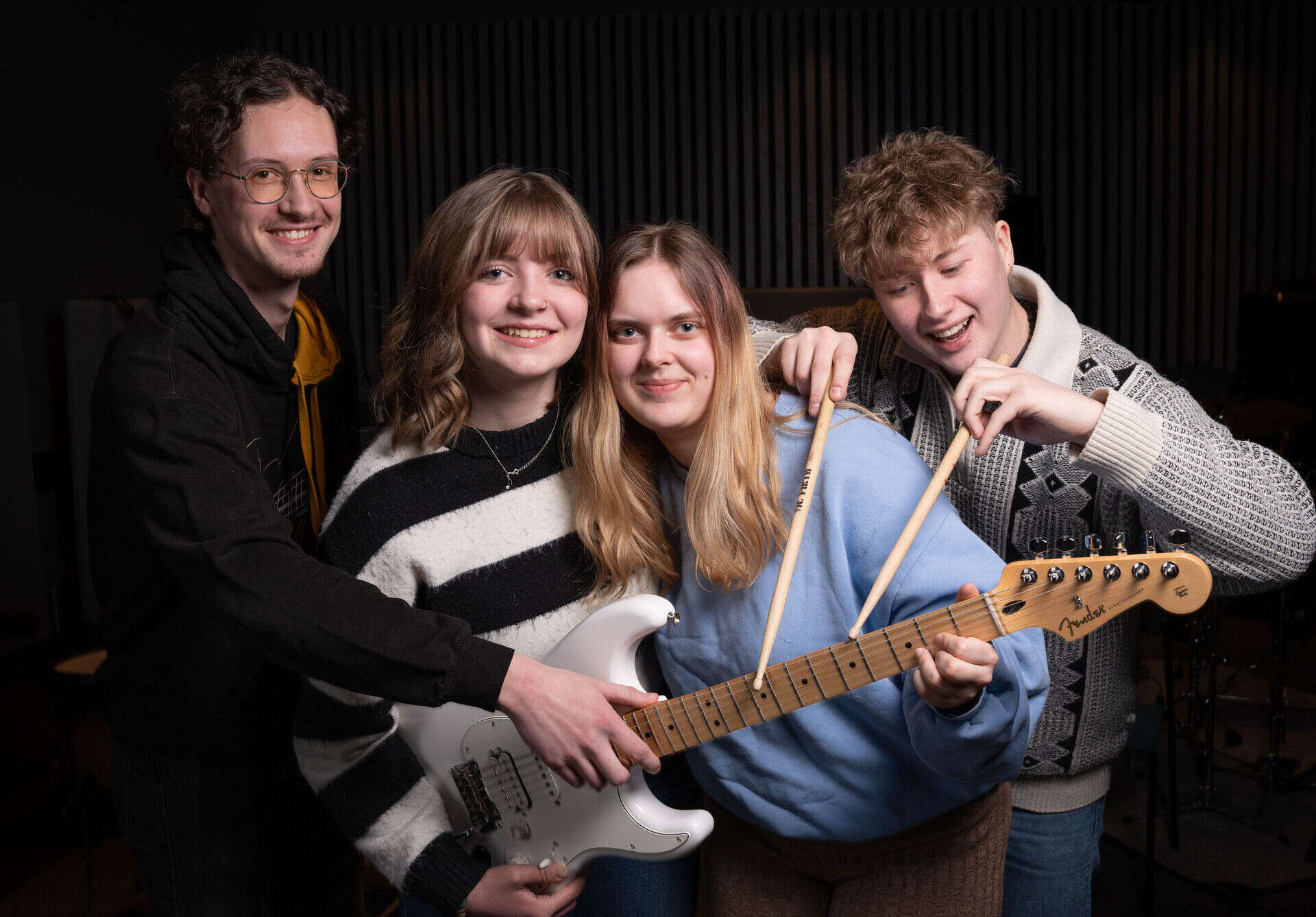 Fire smilende musikkstudenter klynger seg sammen rundt en gitar, og en av studentene trommer med trommestikker på gitarhalsen.