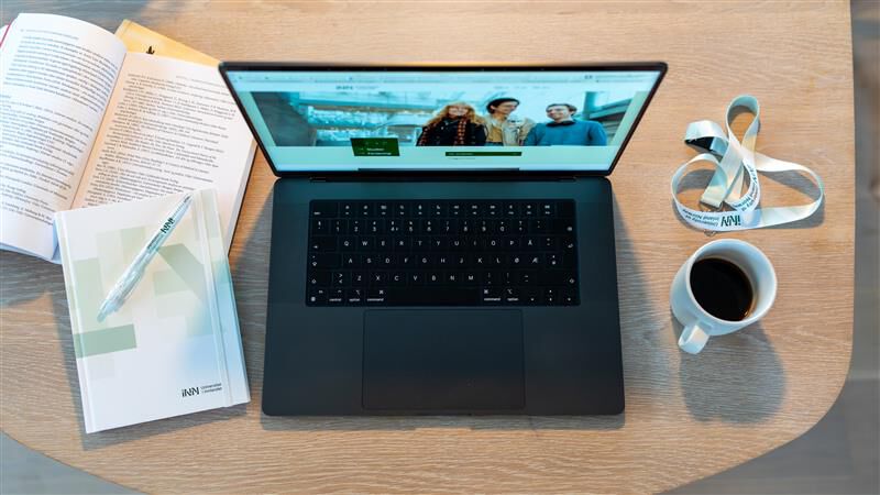 Bilde av laptop, kaffekopp, nøkkelbånd og notatblokk på et trebord