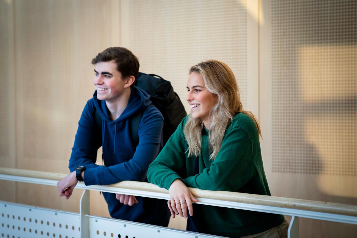 En dame i grønngenser og blondt hår står sammen med en mann med blå hettegenser og mørkt hår står ved et rekkverk og ser utover. 