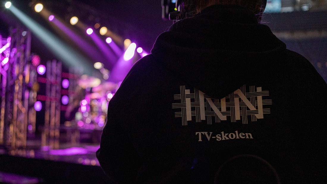 Ryggen til en student ved TV-skolen med svart genser på og logo Høgskolen i Innlandet samt TV-skolen. Ser inn mot scenen det tar opptak ved. 