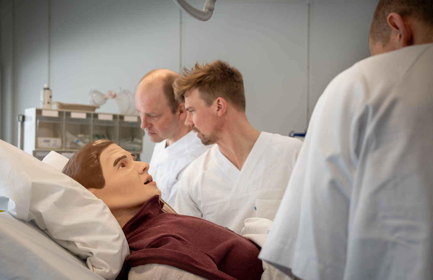 Tre menn i hvite frakker jobbermed ei dukke i ei seng