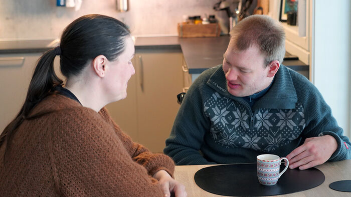 En dame med mørkt hår og brun genser som drikker kaffe med en mann i mørk genser. 