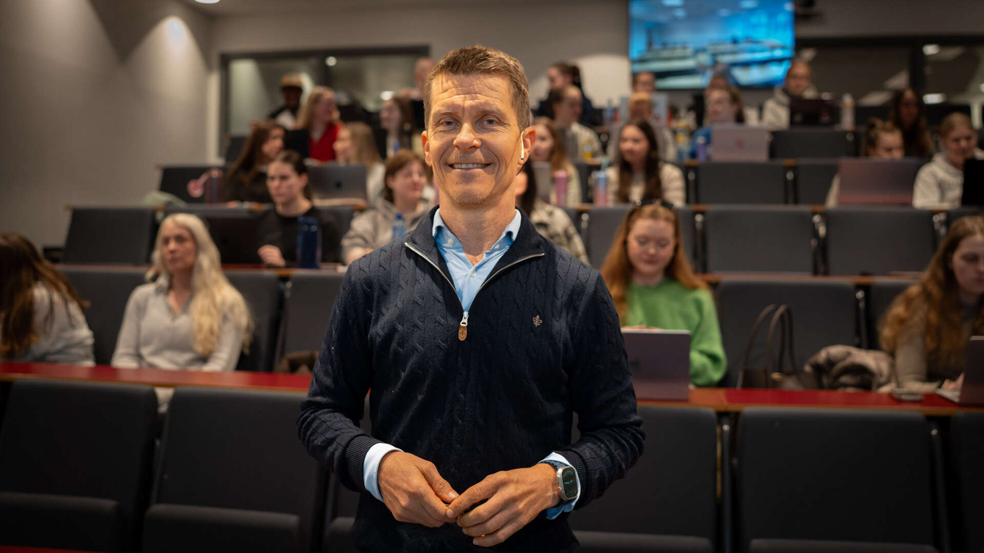 Mann med blå genser står foran en klasse i et auditorium klar for å forelese for dem