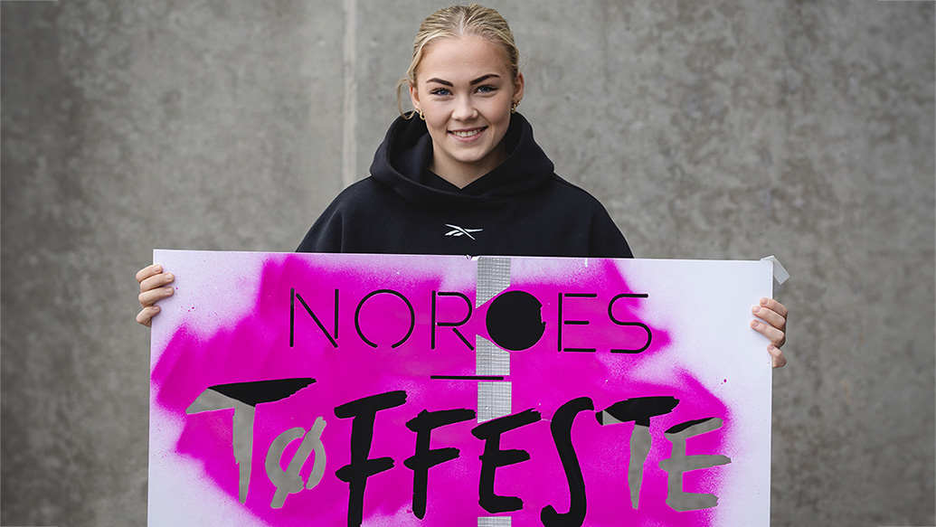 Kvinne i mørke klær som står med en hvit og lilla plakat med teksten Norges tøffeste. 