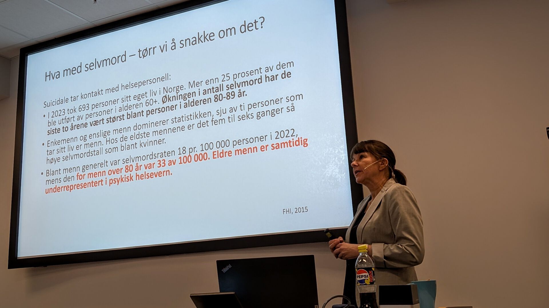 En foredragsholder står foran en projisert presentasjonsslide med tittelen «Hva med selvmord – tør vi å snakke om det?». Sliden viser statistikk og tekst om selvmord blant eldre i Norge, med særlig fokus på menn over 60 år og økningen i selvmordsraten i aldersgruppen 80–89 år. På pulten foran foredragsholderen står en bærbar PC, en brusflaske og noe teknisk utstyr.