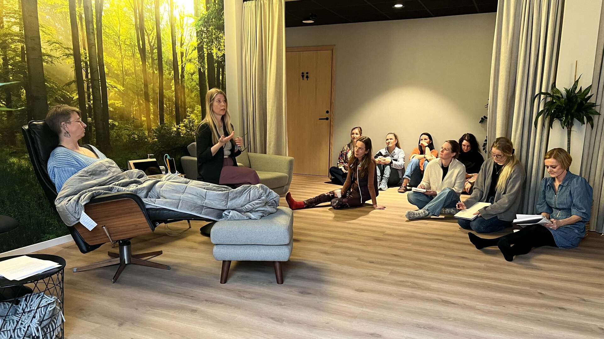 En person sitter i en lenestol, underviser sitter ved siden av, en gruppe studenter sitter på gulvet og følger med og noterer. Stemningsfullt rom med behagelig lys og natur-tapet på veggen.