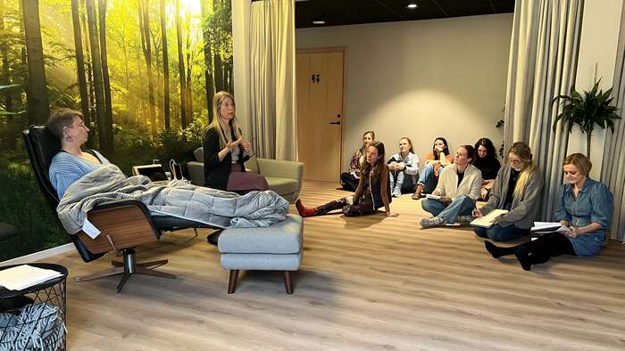 En person sitter i en lenestol, underviser sitter ved siden av, en gruppe studenter sitter på gulvet og følger med og noterer. Stemningsfullt rom med behagelig lys og natur-tapet på veggen.