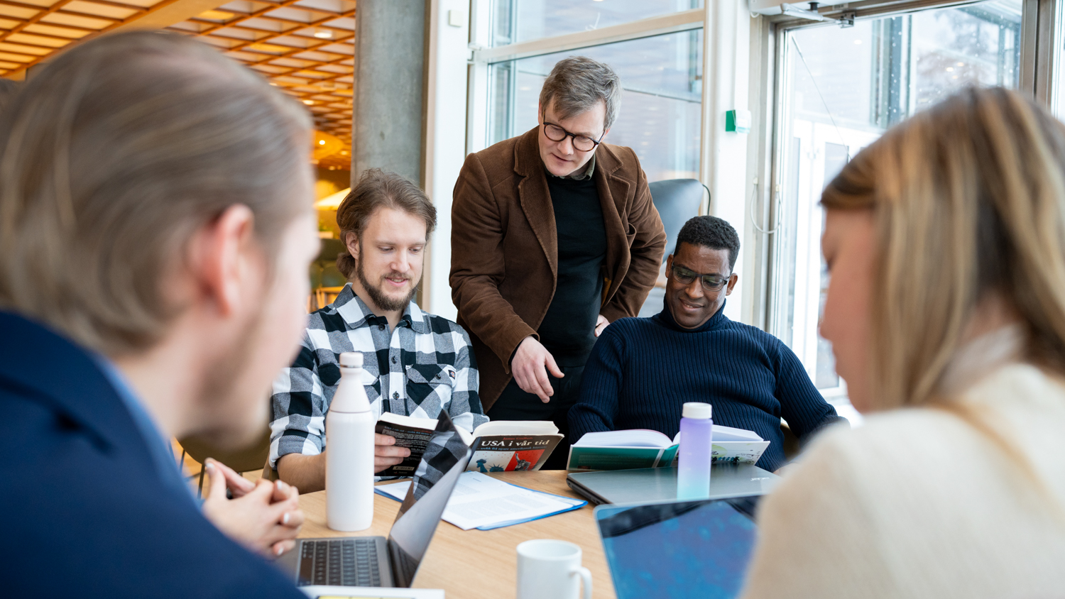 Foreleser Gjermund står og snakker med studenter ved et rundt bord i kantina