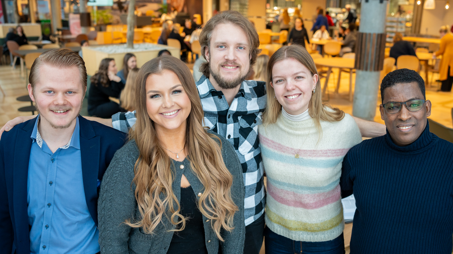 Fem studenter ser i kamera og smiler foran et bord i en kantine.