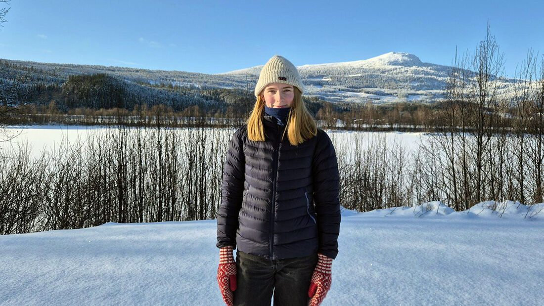 Kvinne i mørke klær som står ute i snøen i strålende vær.