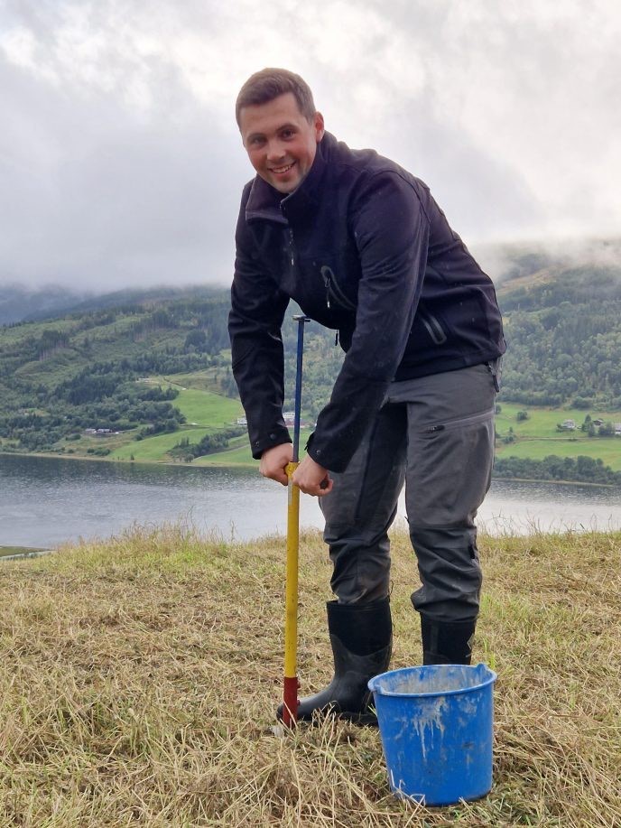Håkon tar en jordprøve. I bakgrunnen kan du se landskapet og fjorden.