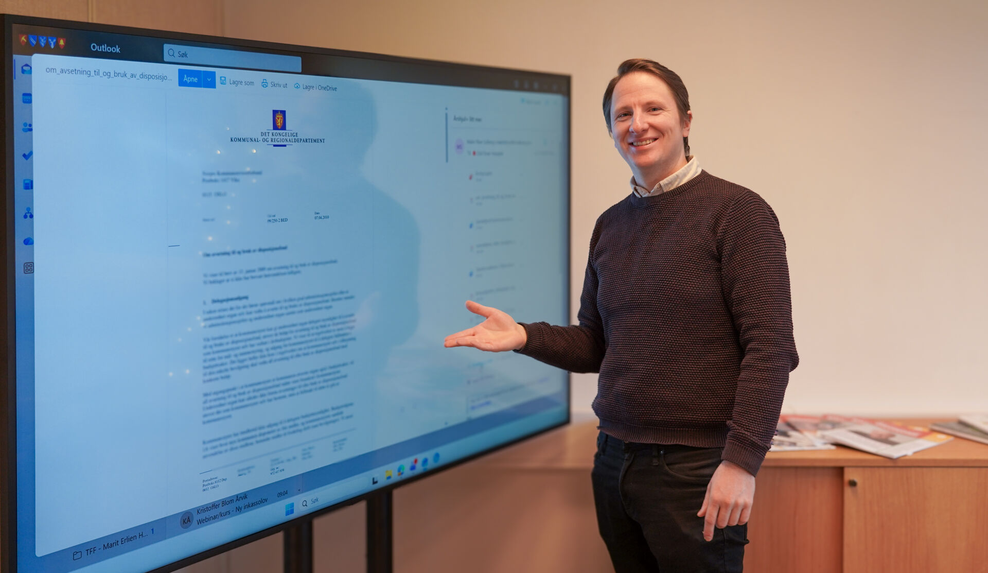 Ung mann med brun genser står og viser fram en stor skjerm på kontoret sitt