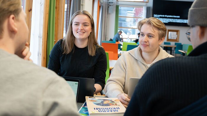 Studenter sitter og snakker sammen ved et bord. En jente og en gutt ser mot kameraet, to gutter med ryggen til. Pensumbøker på bordet. De smiler og ser fornøyde ut.