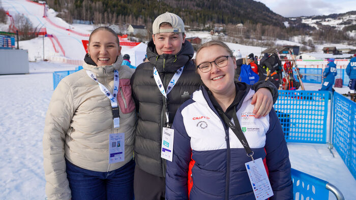 Tre studenter holder rundt hverandre i målområdet i Kvitfjell med alpinister og utforbakken i bakgrunnen