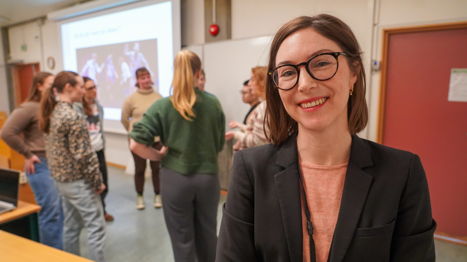 Dame med briller ser i kamera mens studenter jobber i bakgrunnen