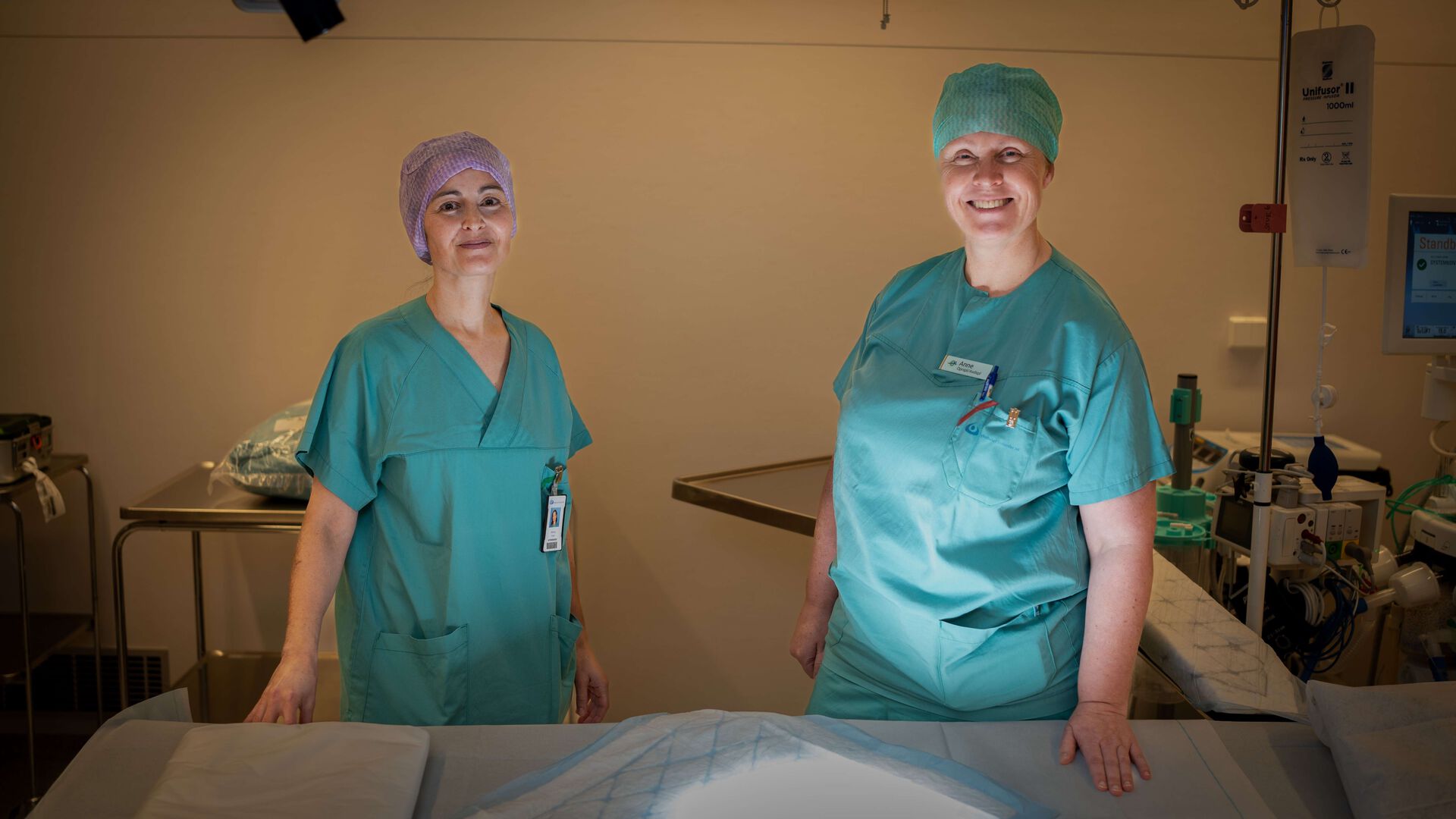 To sykepleiere i grønt står ved en klargjort seng inne på et operasjonssrom. Ser i kamera, lampelys i ansiktet.