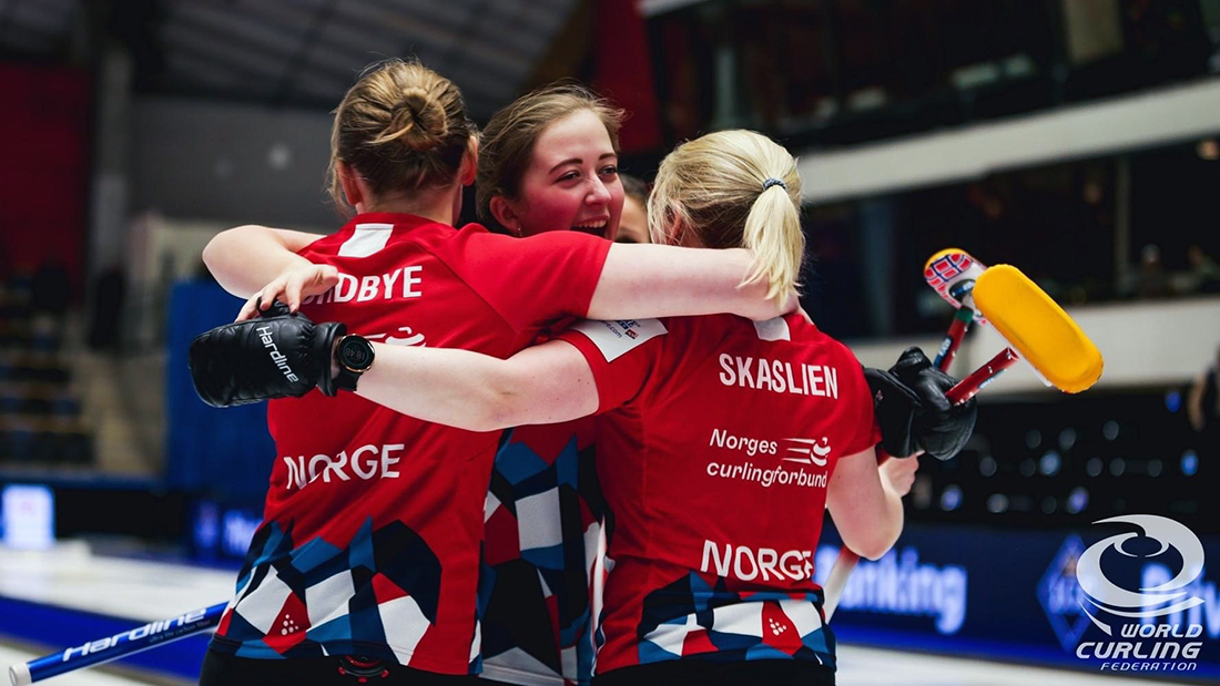 Tre kvinnelige curlingspillere kledd i røde t-skjorter og blå bukser som jubler sammen. 